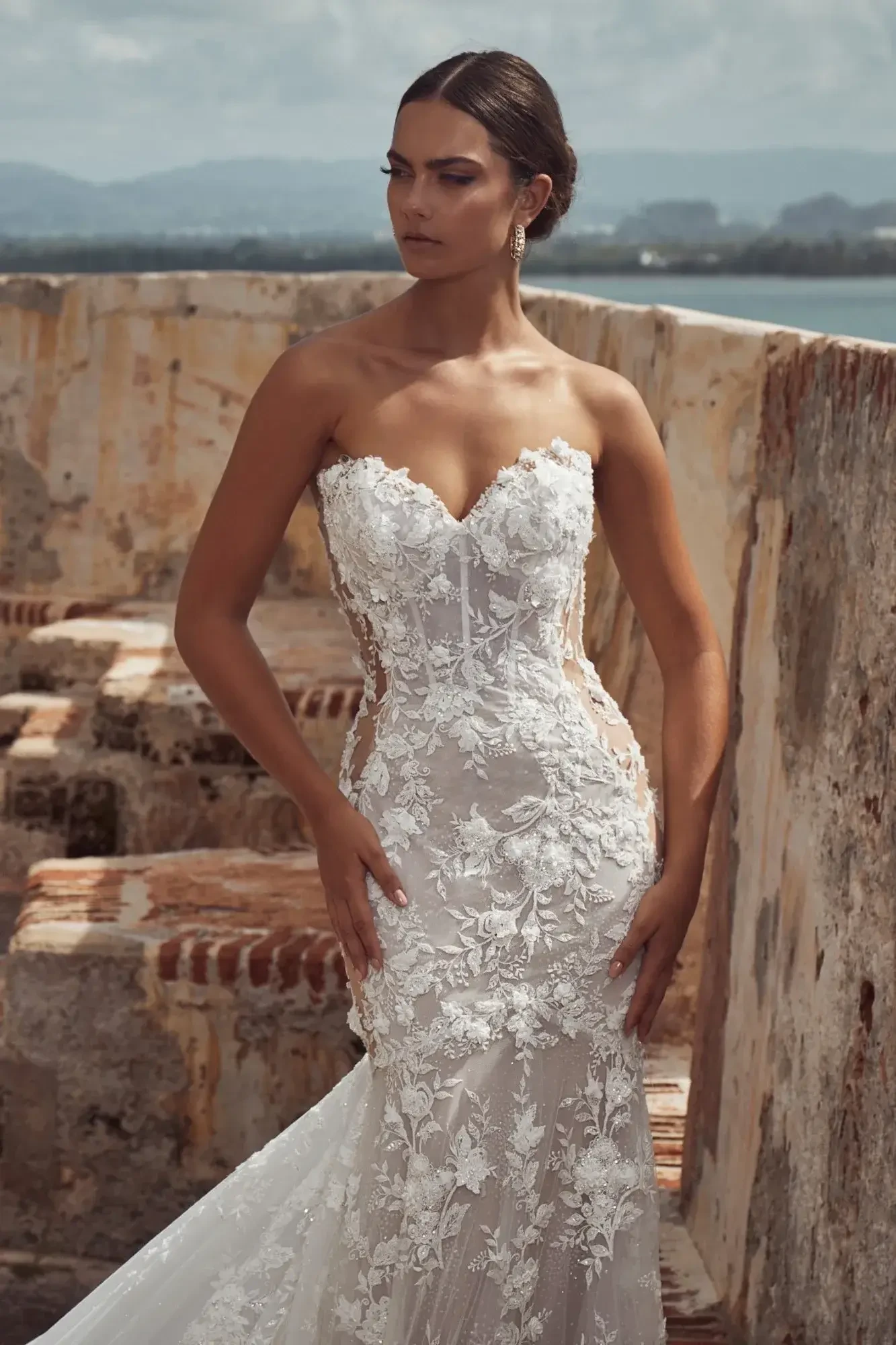A woman wearing a strapless, lace wedding gown with intricate floral designs, standing outdoors near a weathered wall, with a serene expression and the ocean in the background.