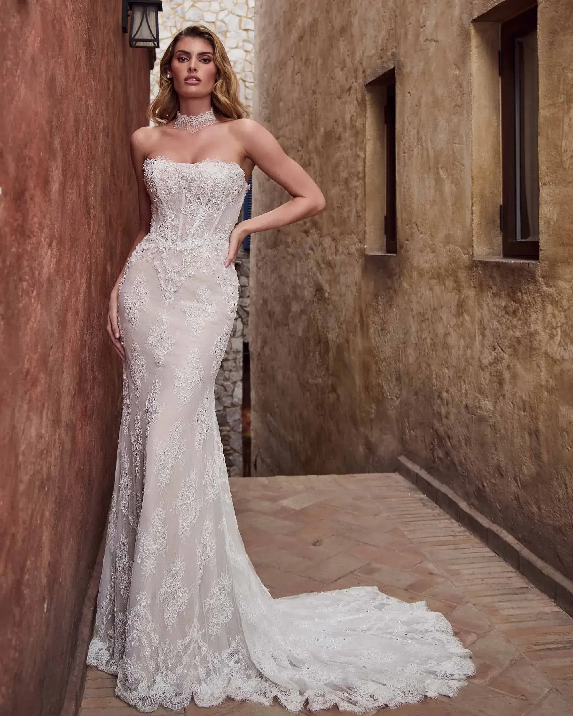 A model is wearing a strapless, fitted wedding gown with intricate lace detailing and a train, standing in a narrow alleyway with textured walls.