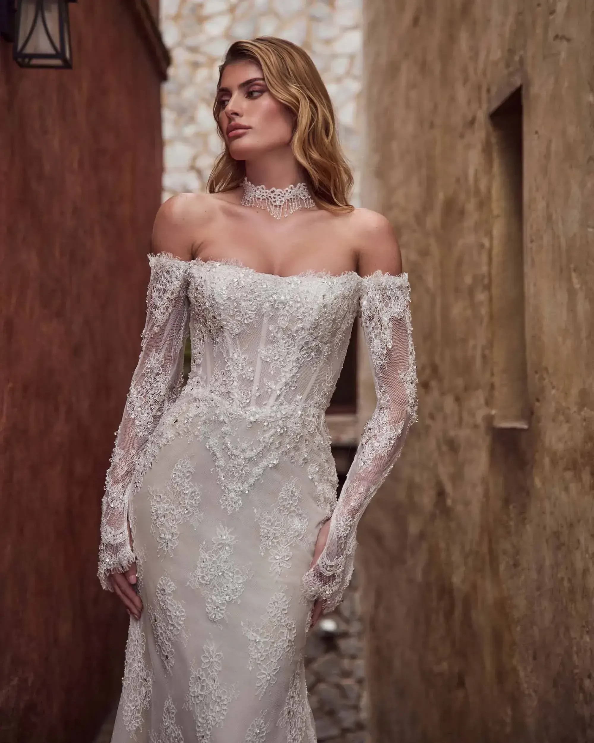 A model wearing an elegant off-the-shoulder wedding dress with intricate lace and beadwork, standing in a narrow alley between textured walls.