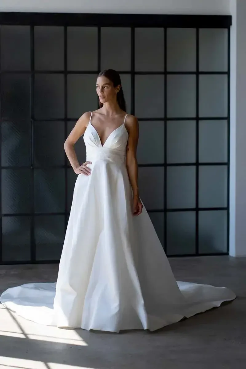 A woman wearing a white wedding dress, standing gracefully in a well-lit room with large windows.