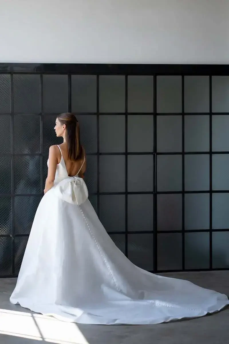 A woman in a simple, elegant white wedding dress with a long train, standing with her back to the camera in a minimalist indoor setting.