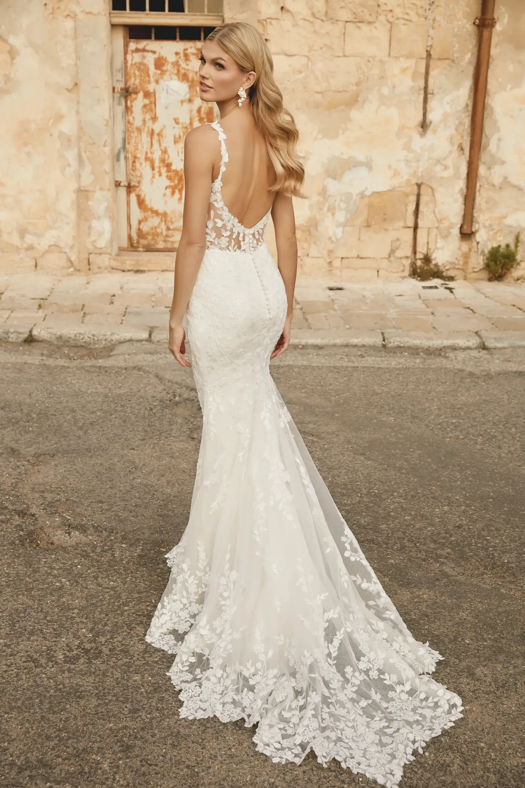 A woman in a backless white wedding dress with intricate lace detailing, standing on a cobblestone street near an aged wall.