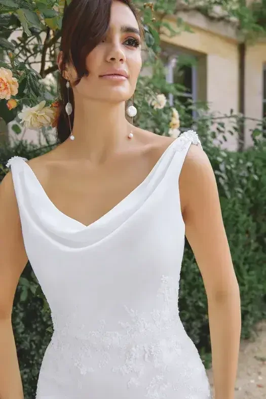 A woman in a white dress with a cowl neckline stands in front of greenery and flowers, looking away from the camera.