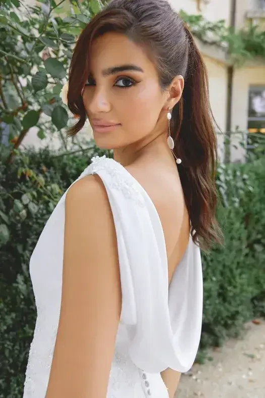 A woman with long, dark hair styled in a ponytail is wearing a white dress with an open back. She is looking over her shoulder at the camera, with greenery visible in the background.