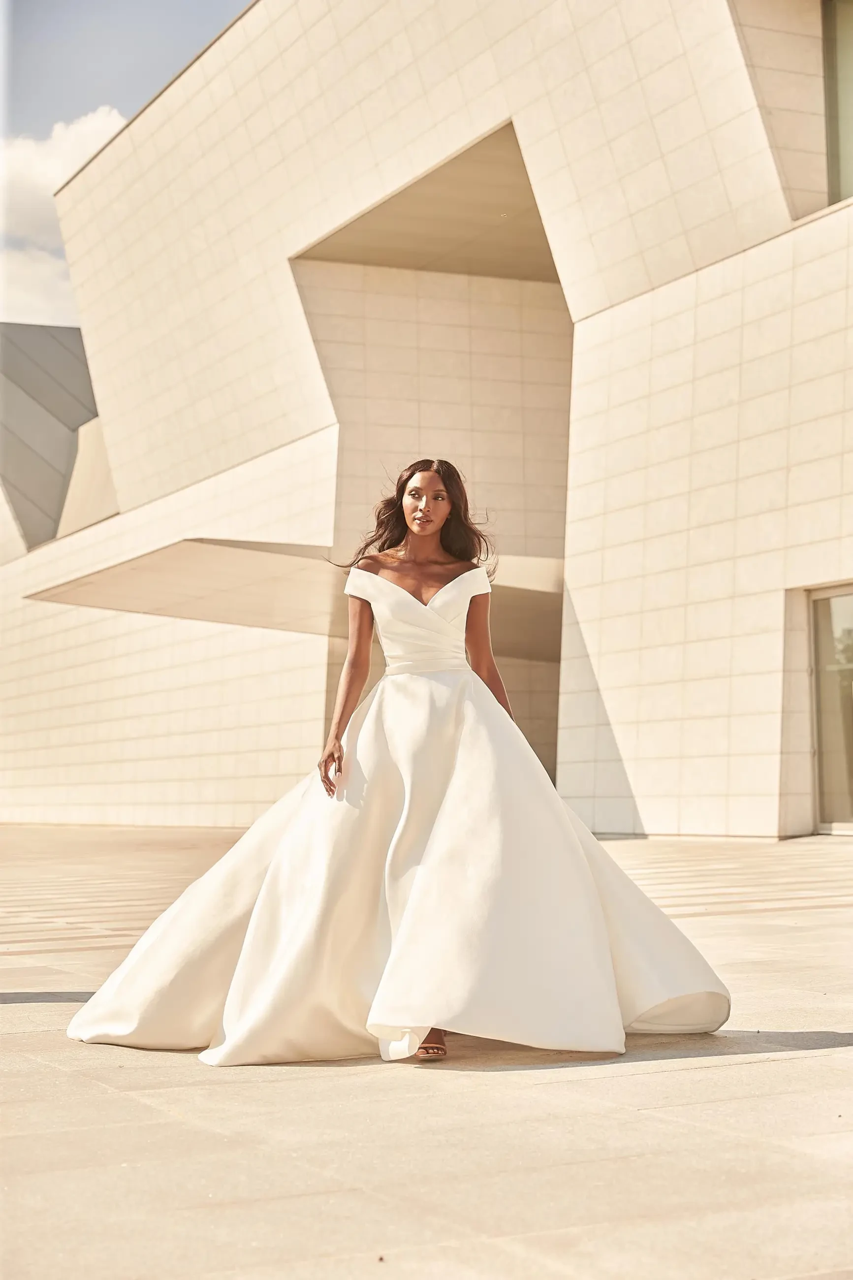 A woman in a flowing white gown stands outdoors in front of a modern architectural structure.