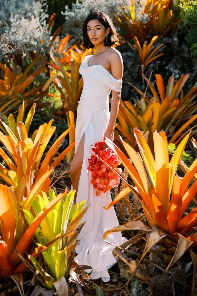 A woman in a white gown stands among vibrant orange and green plants, holding a bouquet of red flowers.