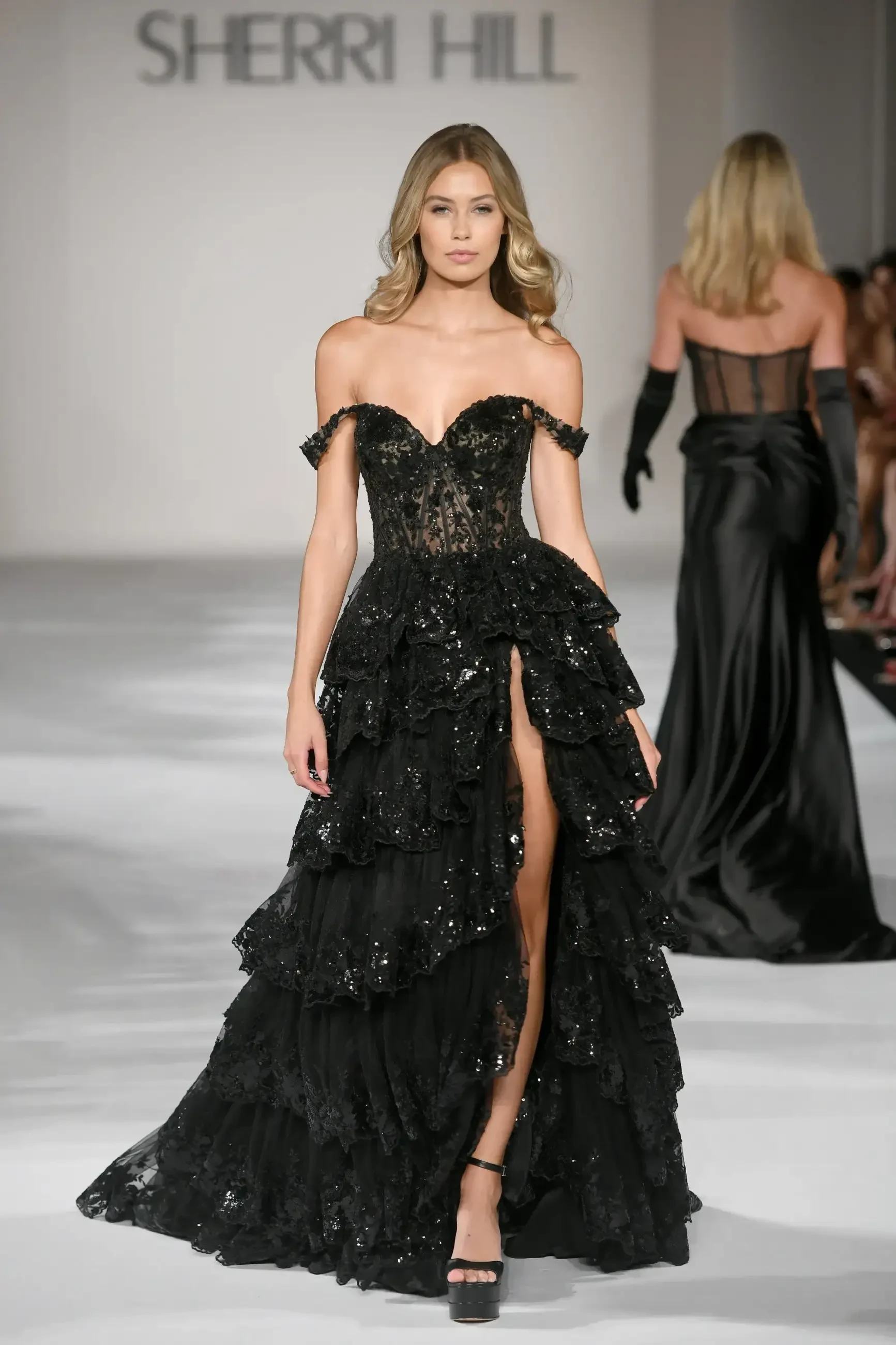 A model walks the runway wearing a black strapless gown with layered ruffles and a high slit, during a fashion show for Sherri Hill.