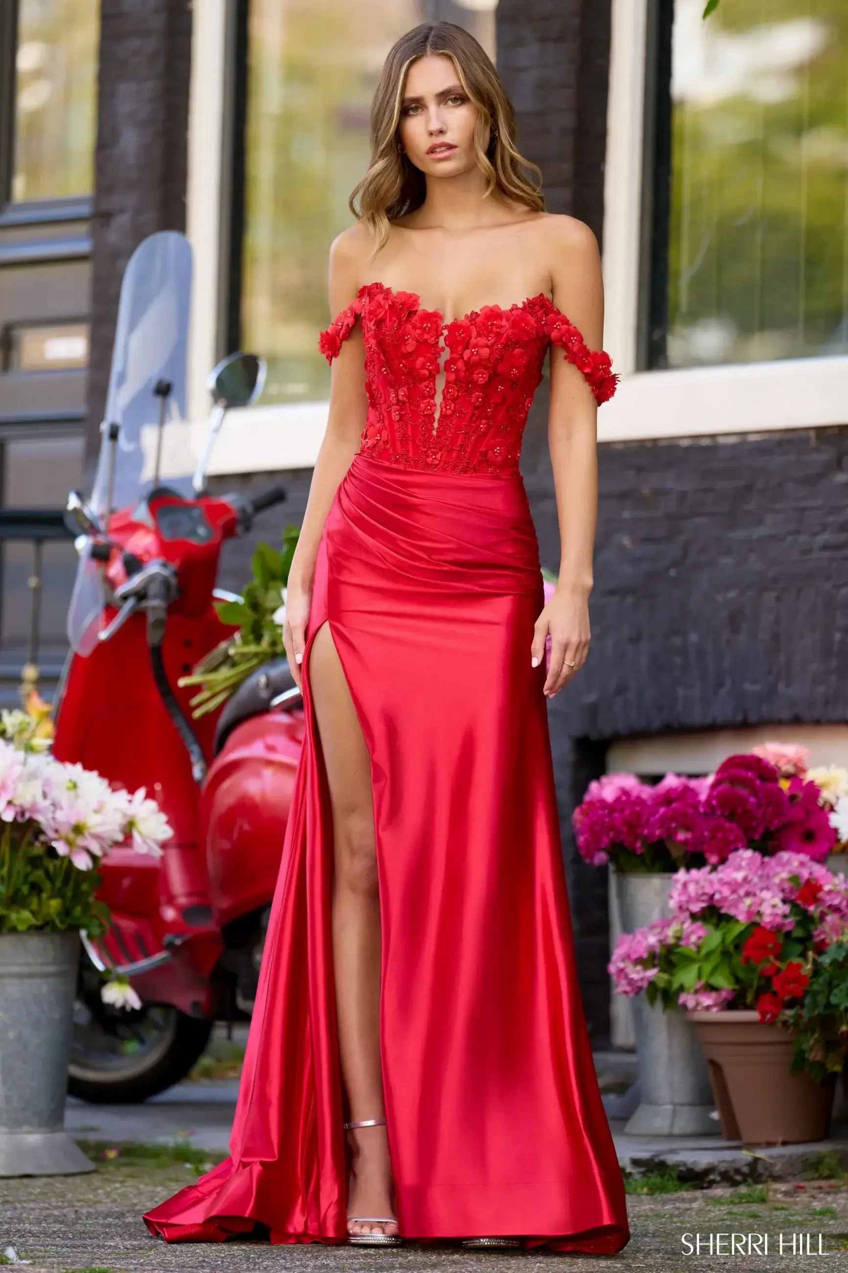 A woman wearing a red off-the-shoulder evening gown with floral embellishments and a thigh-high slit, standing in front of a background with flowers and a vintage scooter.
