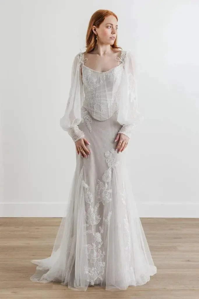 A woman in a white, vintage-inspired dress with long sleeves and floral lace details, standing against a plain background.