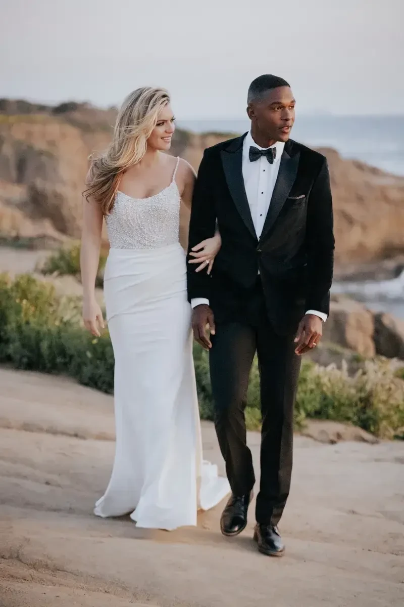 A couple walking together along a rocky coastline, with the ocean in the background. The woman is wearing a white dress and the man is dressed in a black tuxedo with a bow tie.