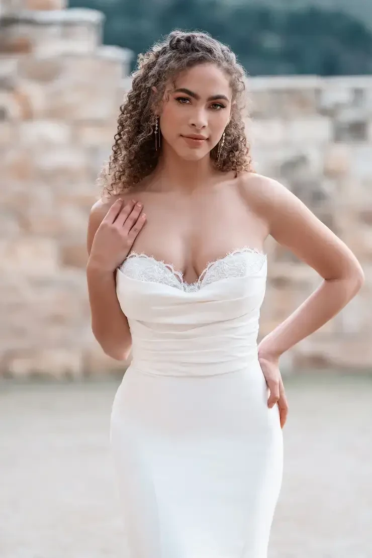 A young woman with curly hair wearing a white strapless gown, standing in front of a stone wall.