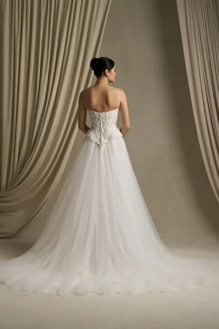 A woman in a strapless white wedding gown, viewed from the back, with a flowing skirt and lace detailing. The background features draped fabric.