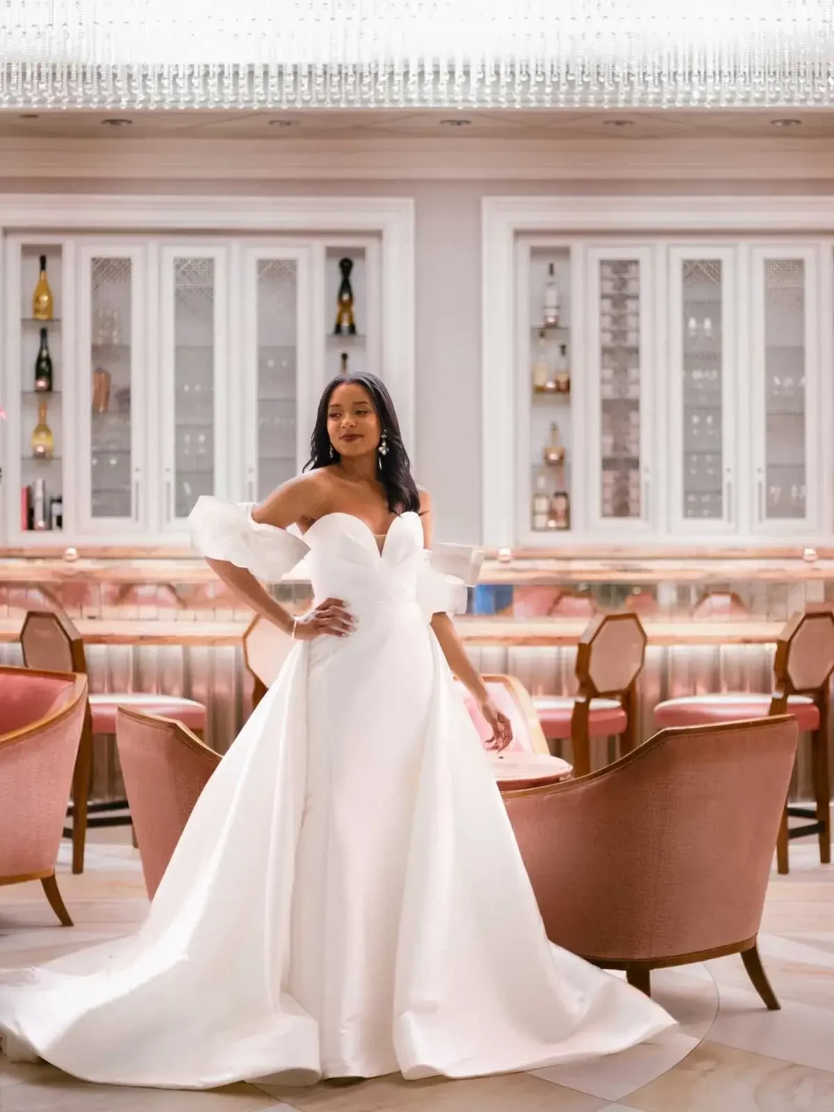 A woman wearing a white off-the-shoulder gown stands elegantly in a chic, well-lit bar with pink upholstered chairs and shelves displaying bottles.