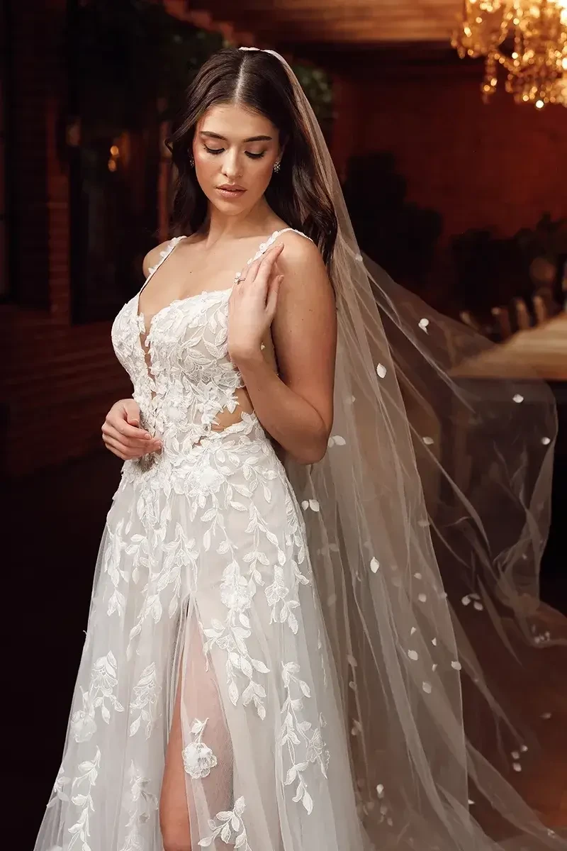 A model wearing a wedding dress with floral embroidery and a long veil, posing in an elegantly decorated venue.