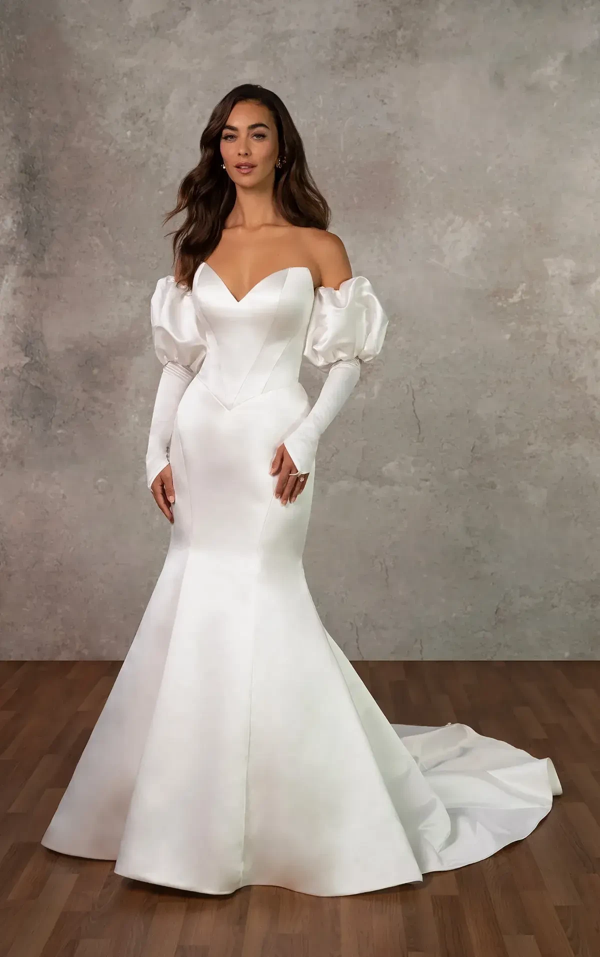 A woman in a strapless white wedding gown with puffed sleeves and a mermaid silhouette, standing on a wooden floor against a gray backdrop.