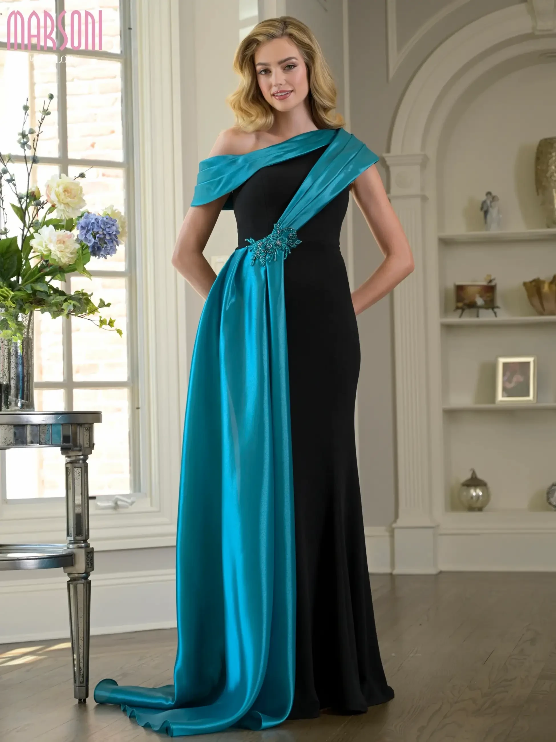 A woman wearing an elegant black and turquoise dress, standing in a well-lit room with decorative plants and floral arrangements.