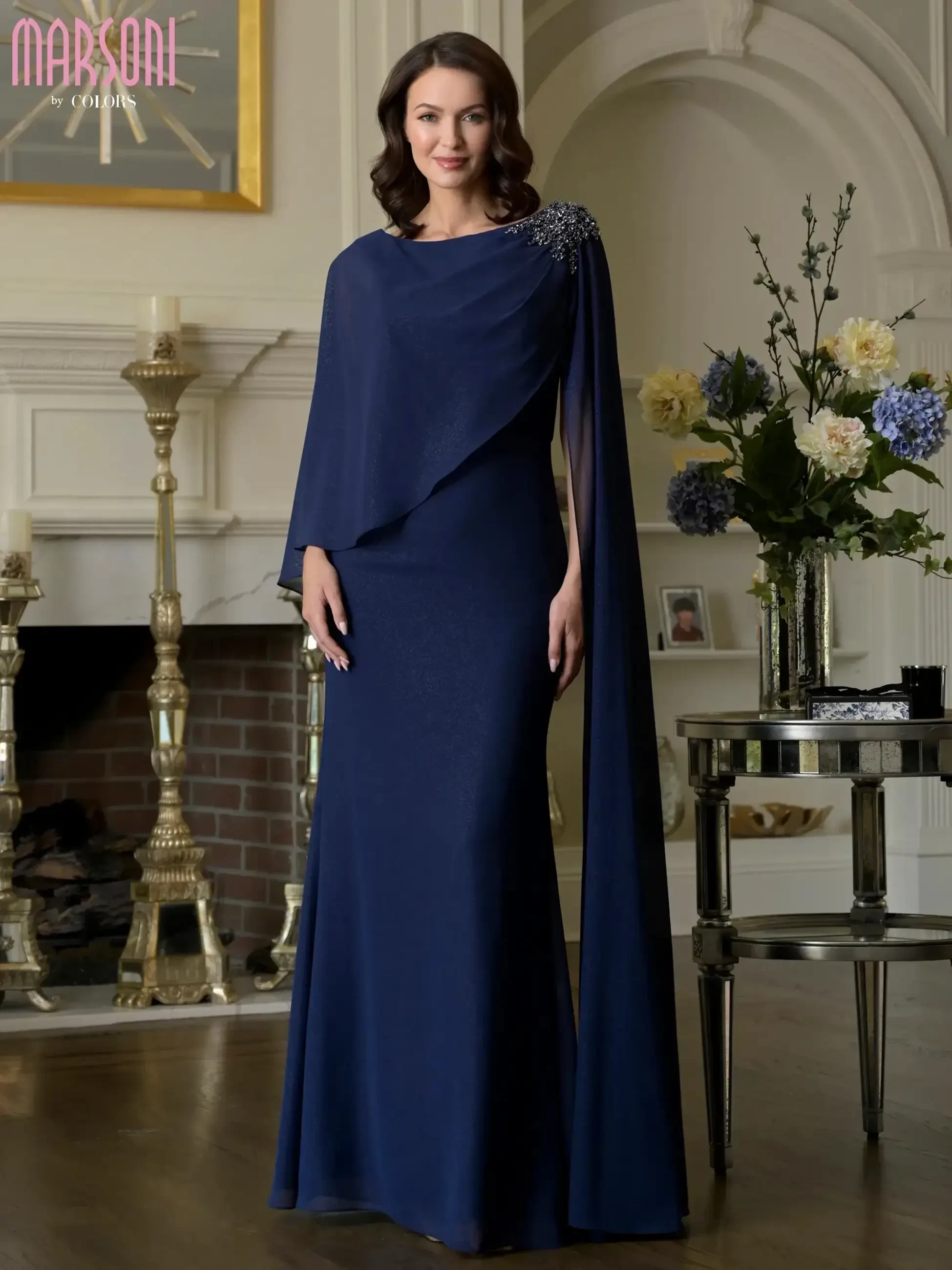 A woman wearing a navy blue evening gown with a cape-like design, standing elegantly in a stylish room with decorative items in the background.