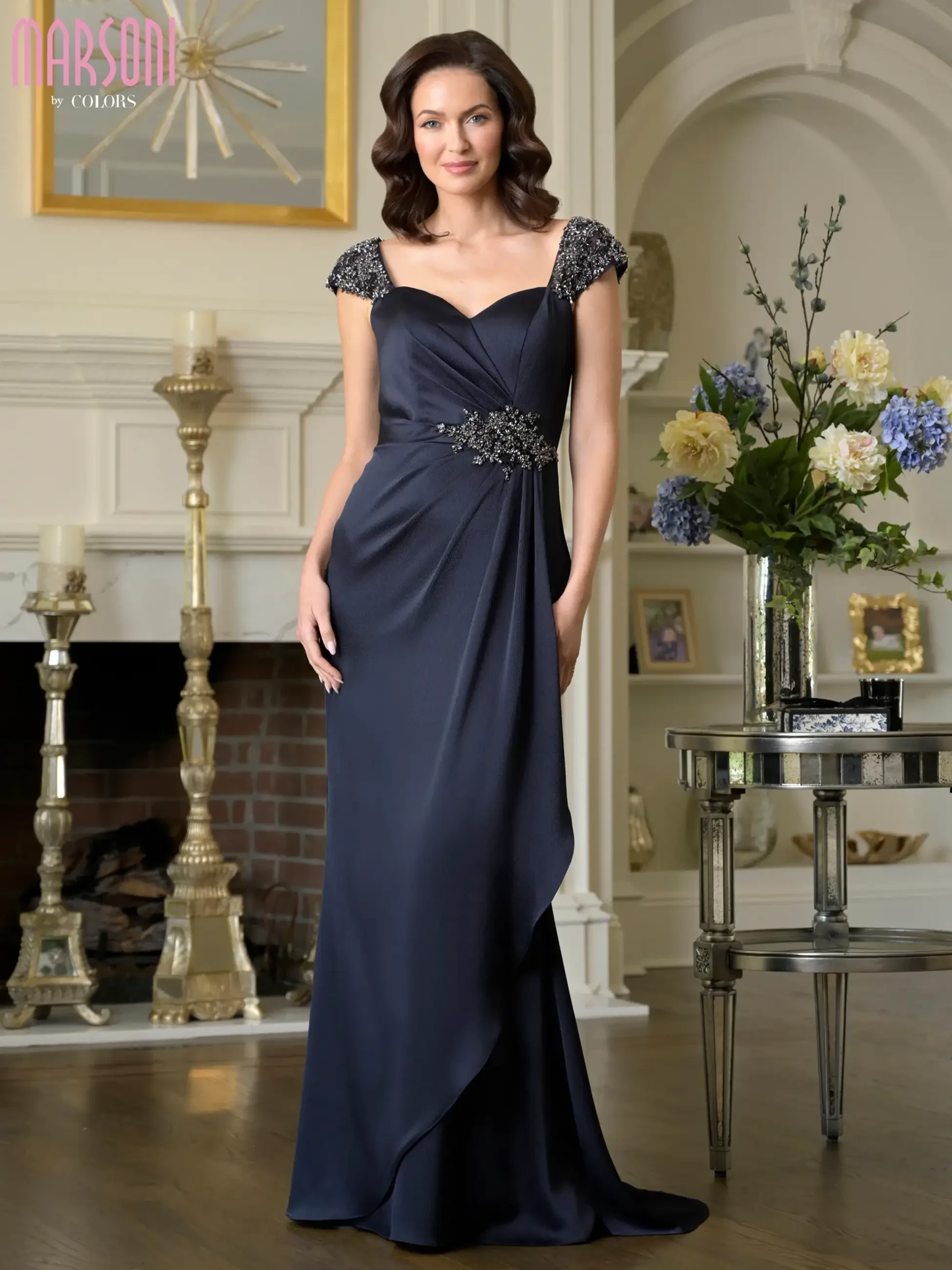 A woman in an elegant navy blue evening gown with embellished shoulders stands in a tastefully decorated room featuring a fireplace and a floral arrangement.