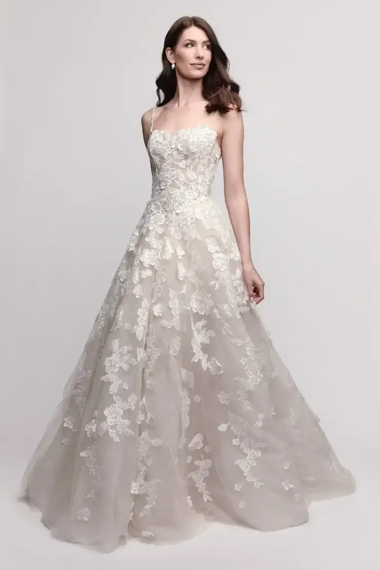 A woman wearing a beautiful, flowing wedding gown with a floral lace design and a fitted bodice. The dress has thin spaghetti straps and is complemented by delicate detailing throughout the skirt.