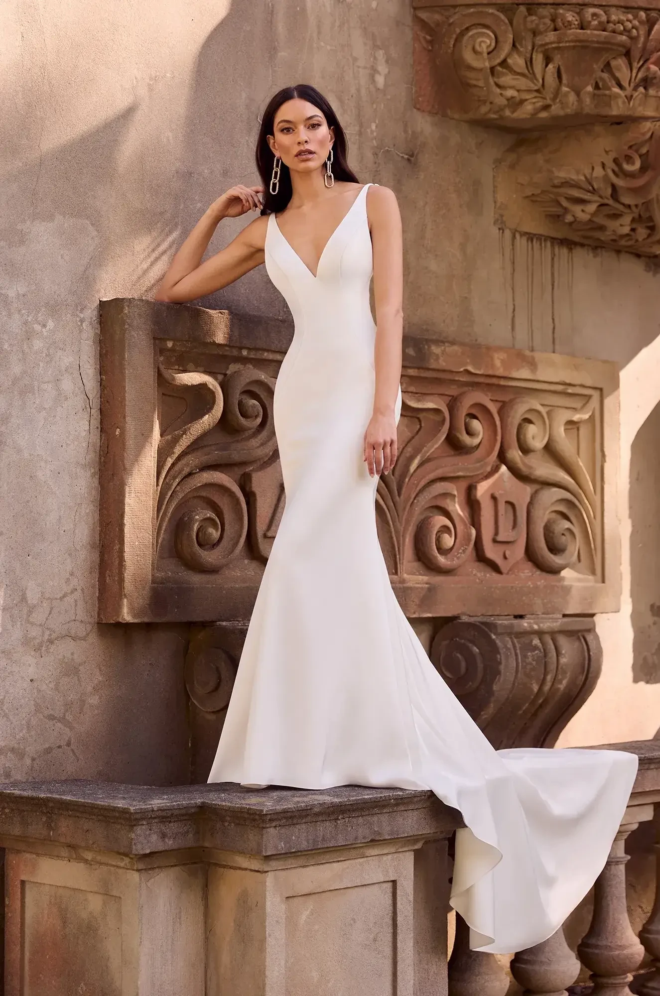 A model poses in a sleek white gown with a fitted silhouette and a mermaid train, standing on a stone ledge against an ornate wall backdrop.