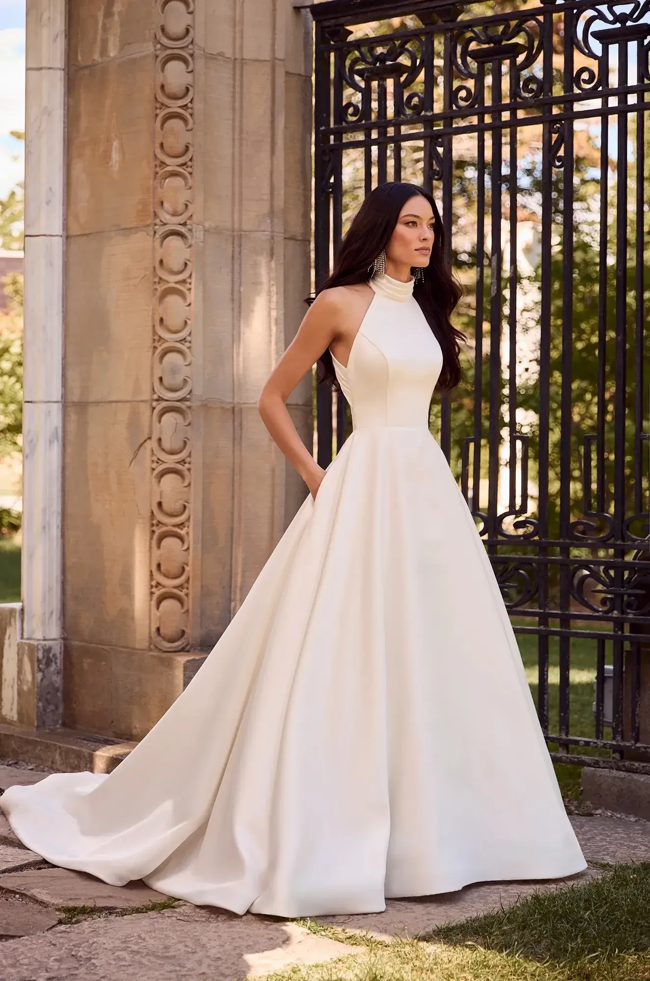 A woman in a sleeveless white gown stands outside near a decorative iron gate, with her hands in the pockets of the dress. The background features greenery and trees.