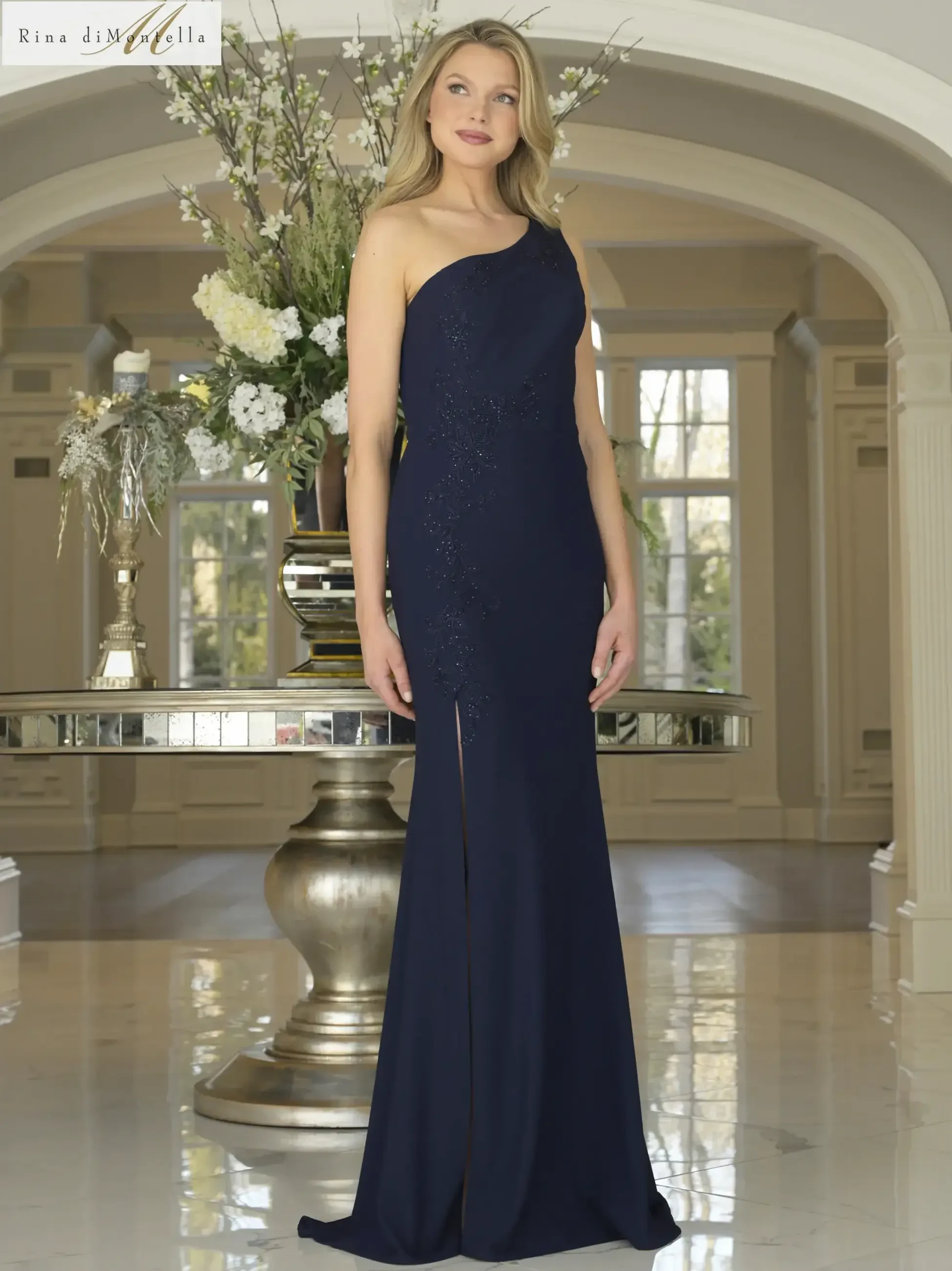 A model wearing a navy blue one-shoulder evening gown with a slit, standing in an elegant interior setting with floral arrangements.