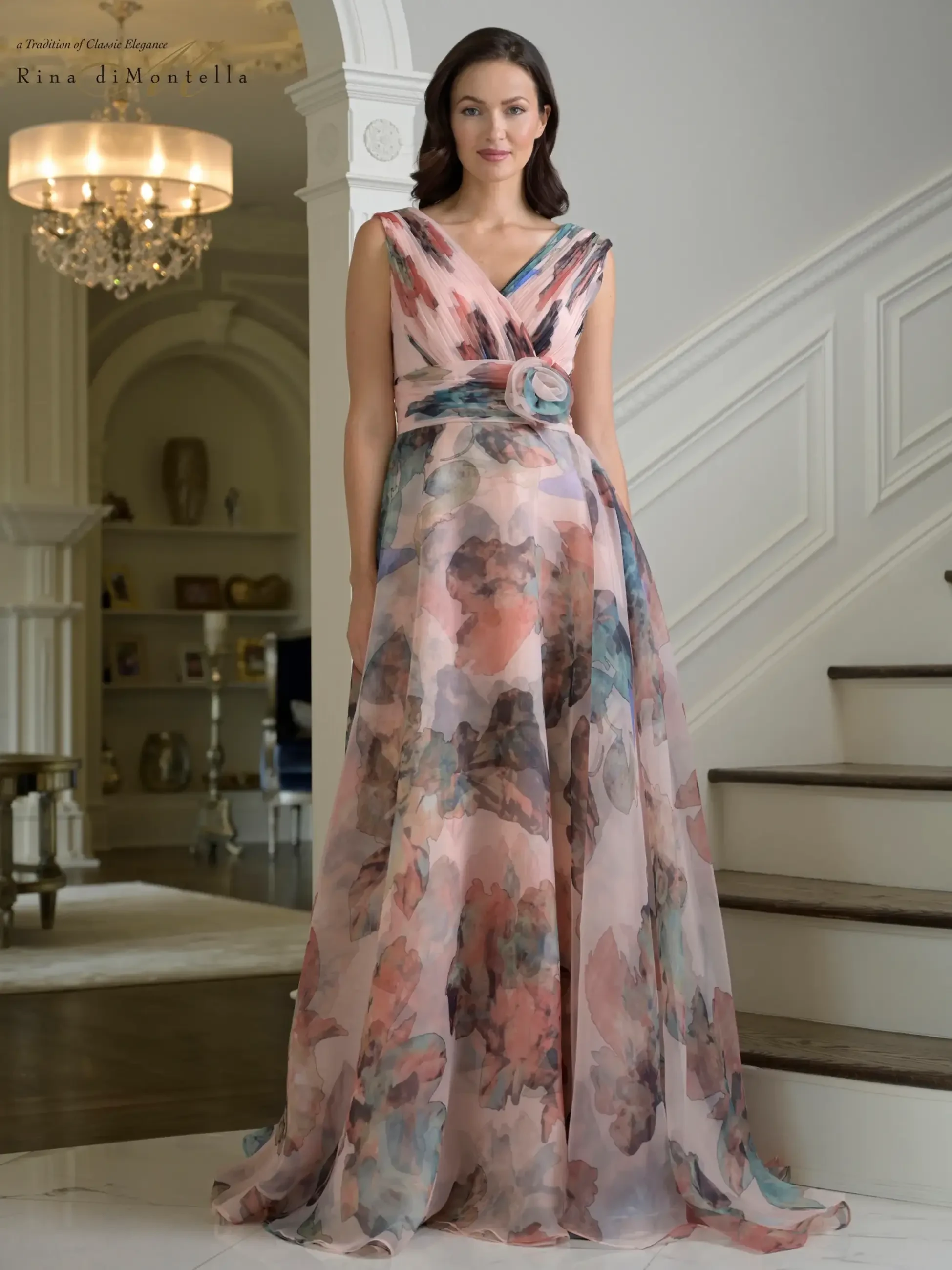 A model wearing a flowing gown with a floral print, standing on a staircase in an elegant interior.